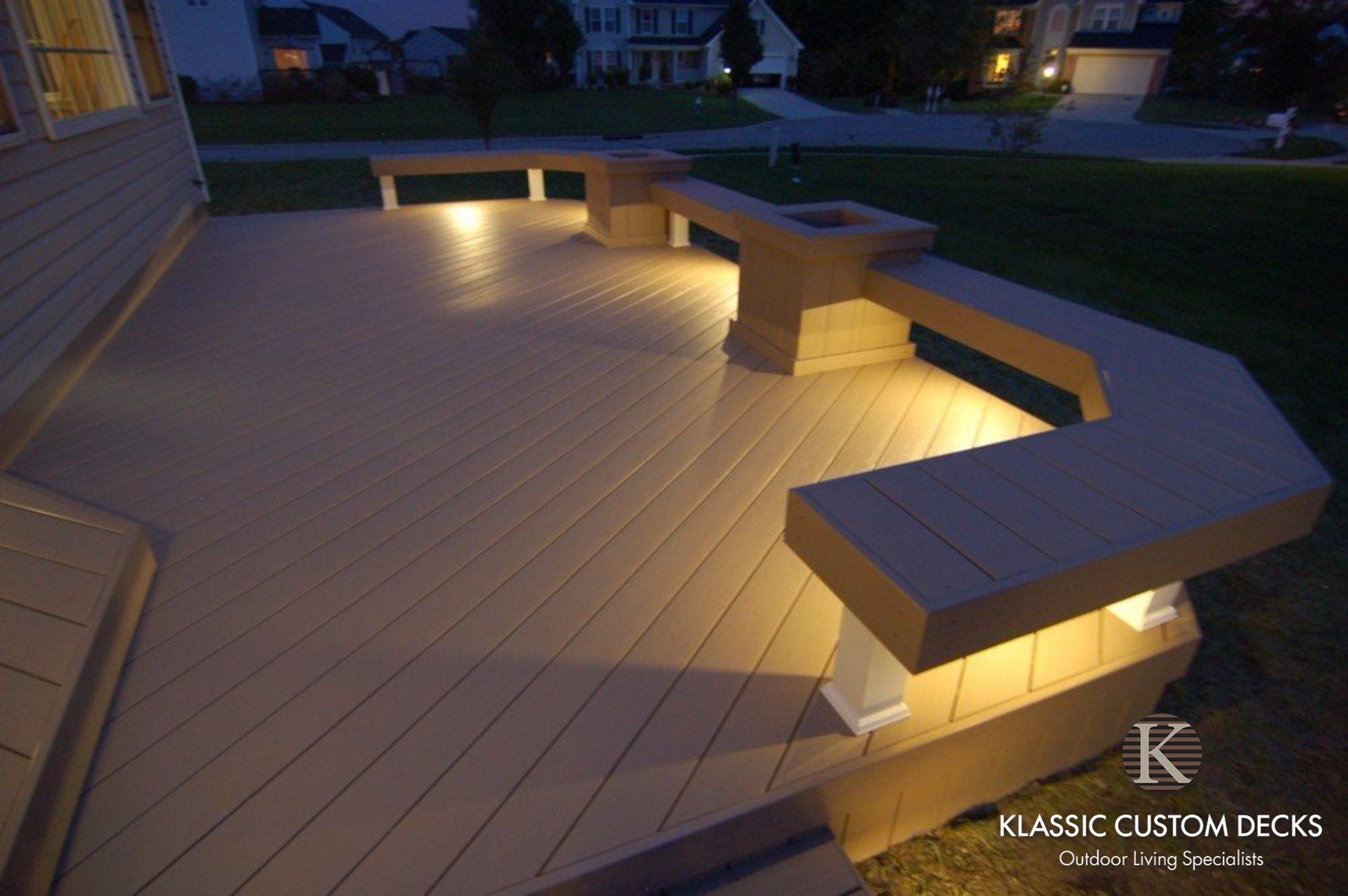 Illuminated wooden deck with built-in benches at dusk, overlooking a yard and houses.