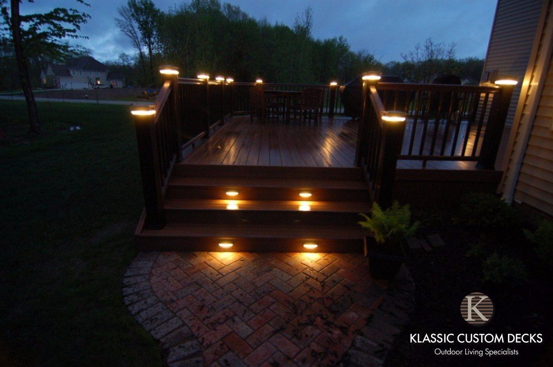 Wooden deck stairs with warm step lights at dusk, leading to a patio with railings and brick pavers.