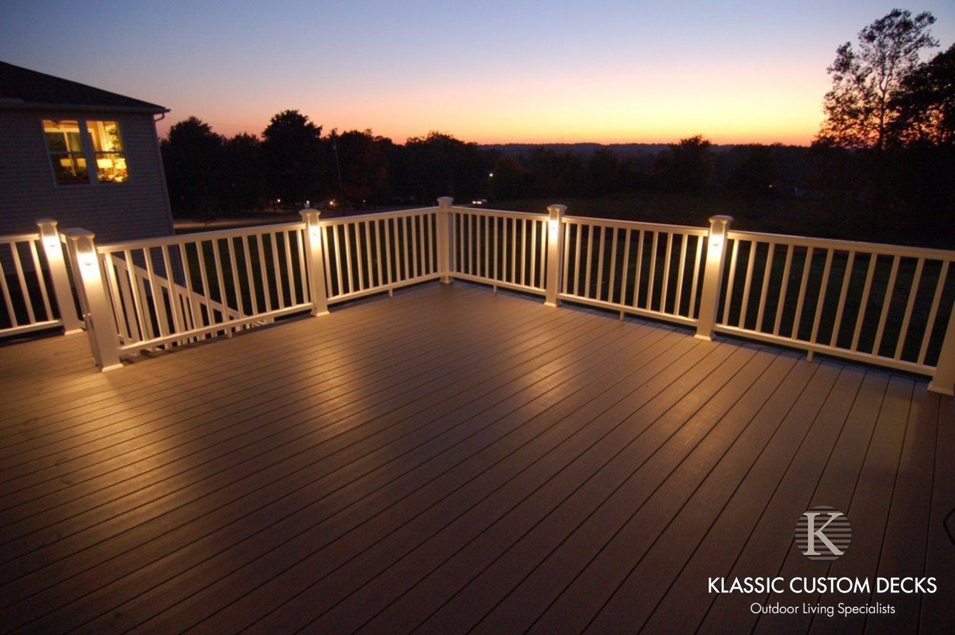 Spacious wooden deck at dusk with white railings and warm lights overlooking a dark landscape.