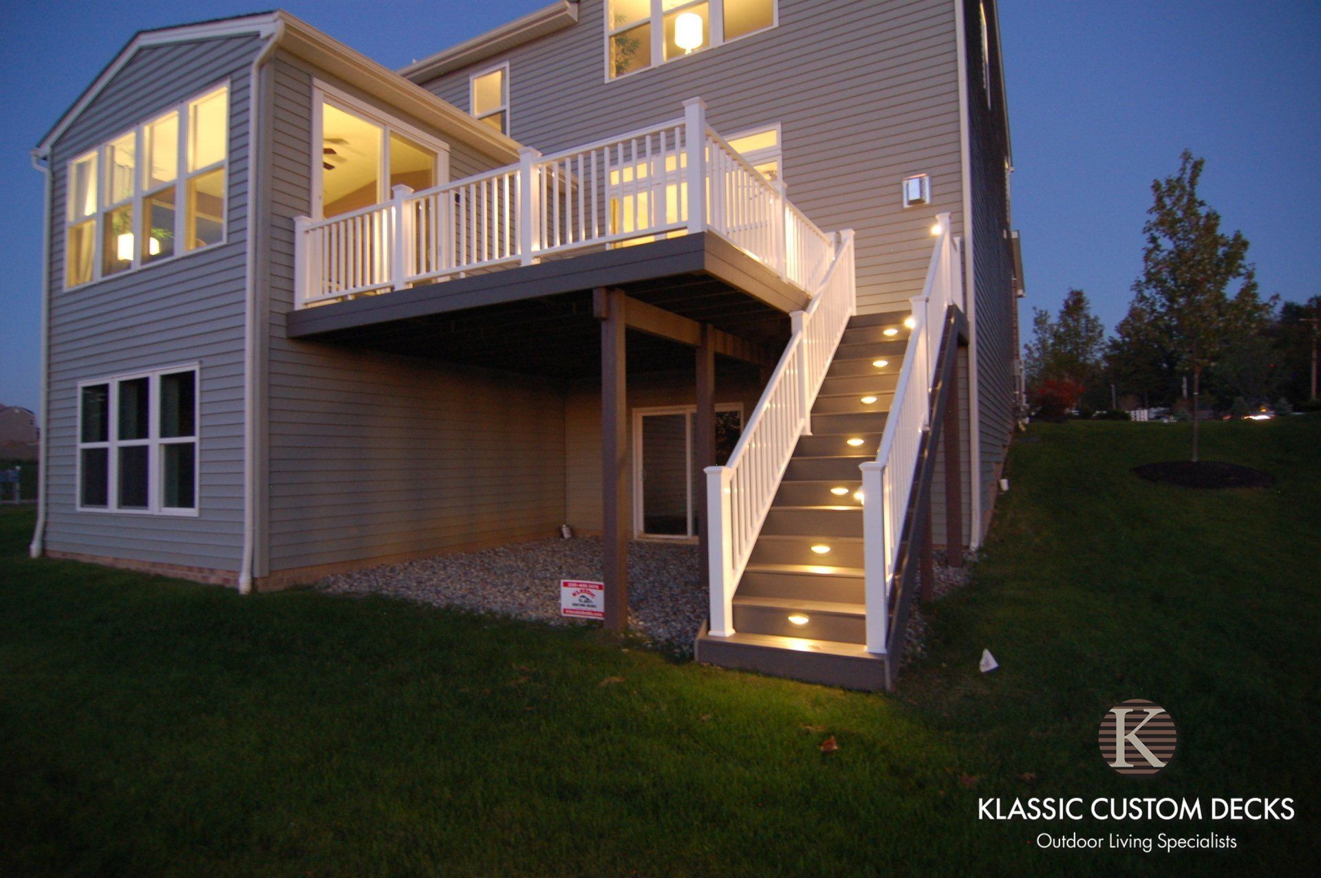Backyard deck and staircase at dusk on a beige house with lit windows and white railings