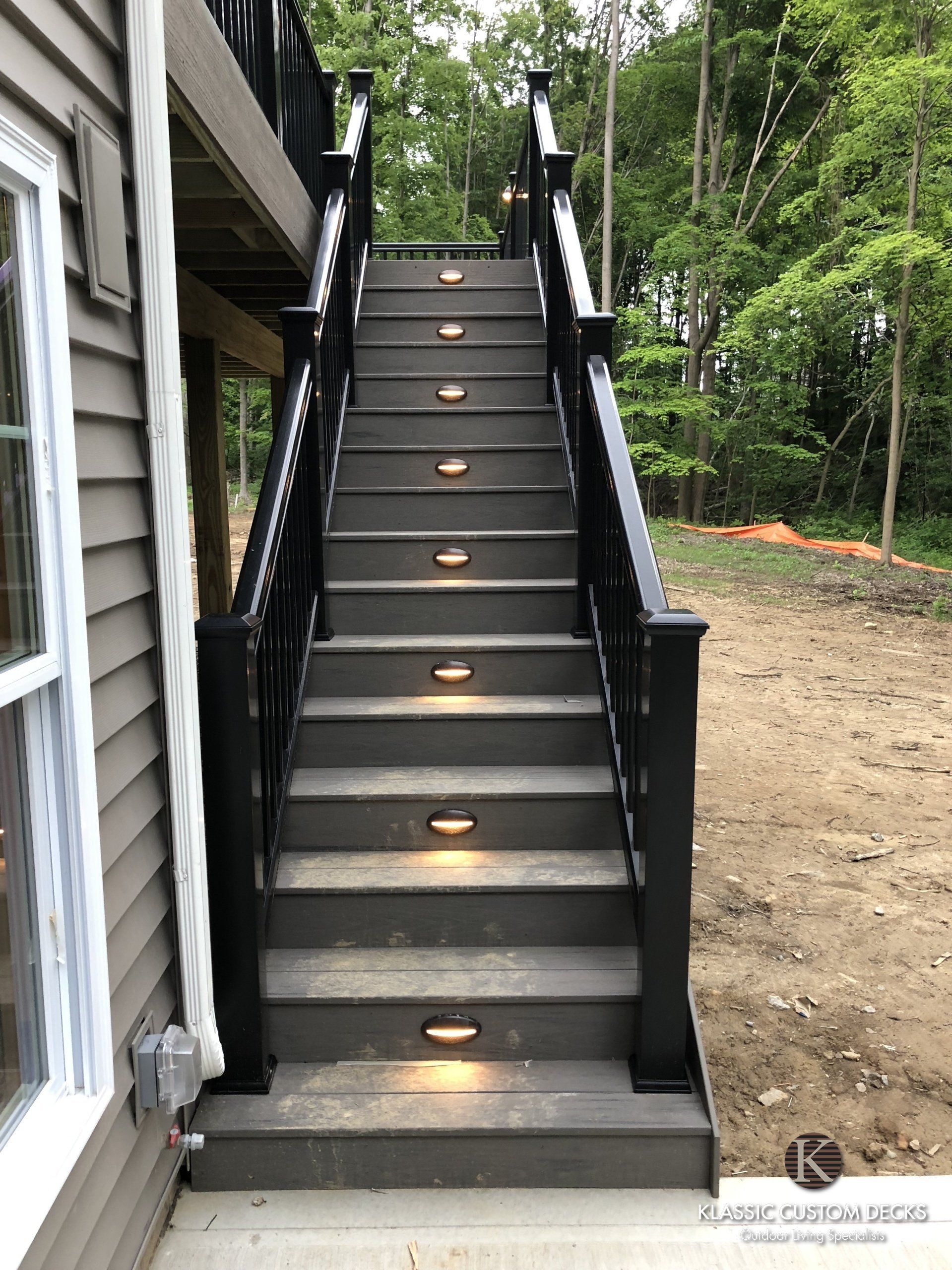 Black outdoor staircase with step lights beside a house, leading up to a wooded yard.