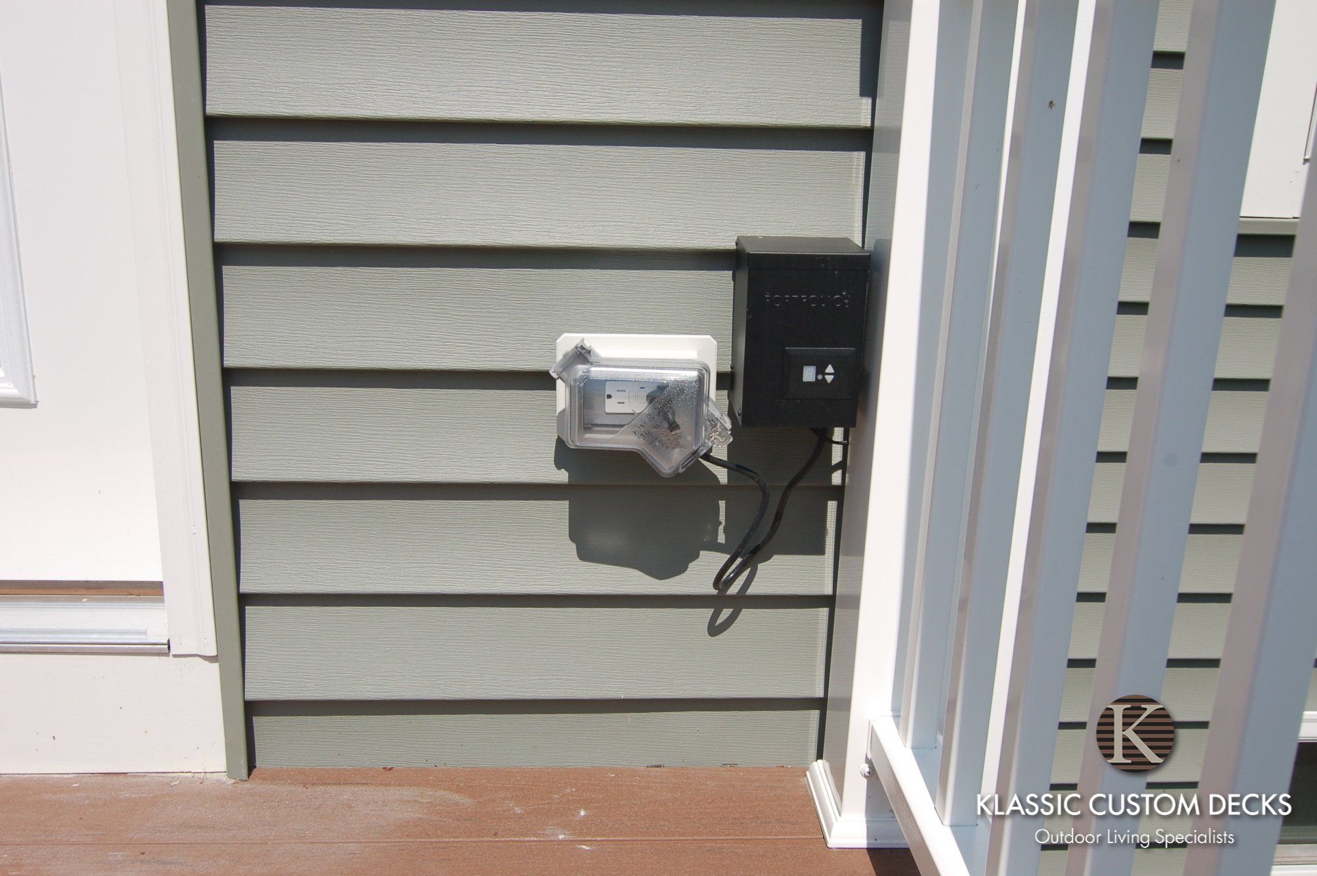 Wall-mounted electric meter and black utility box on gray house siding beside a porch railing.