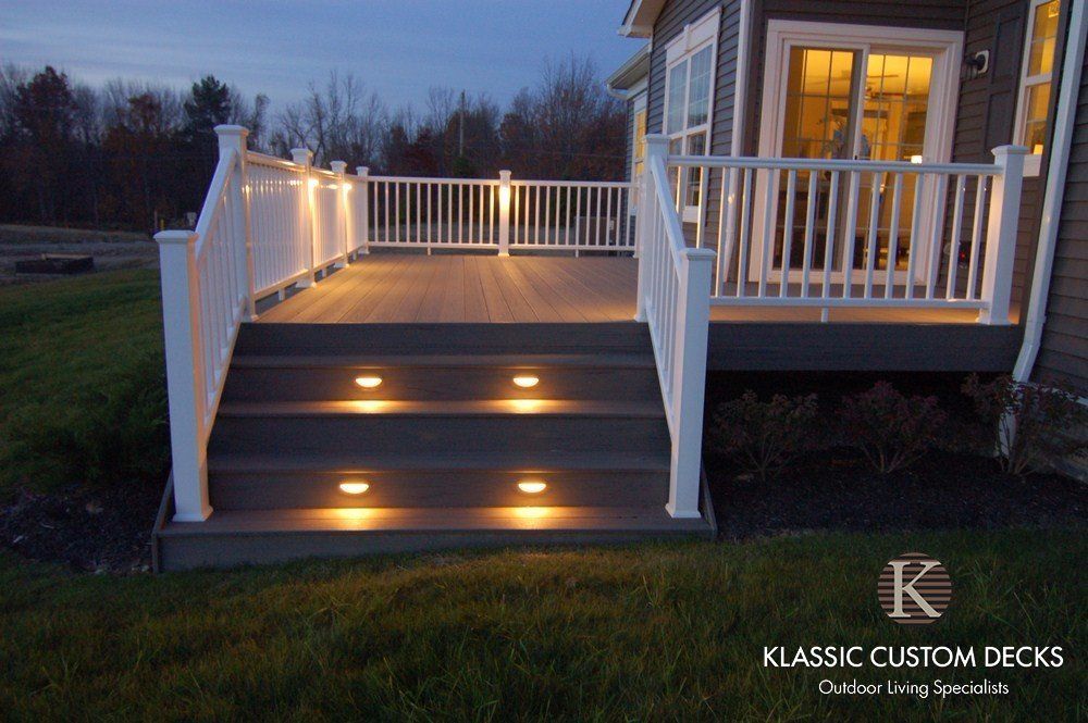 Illuminated wooden deck stairs with white railings at dusk, leading to a lit porch.