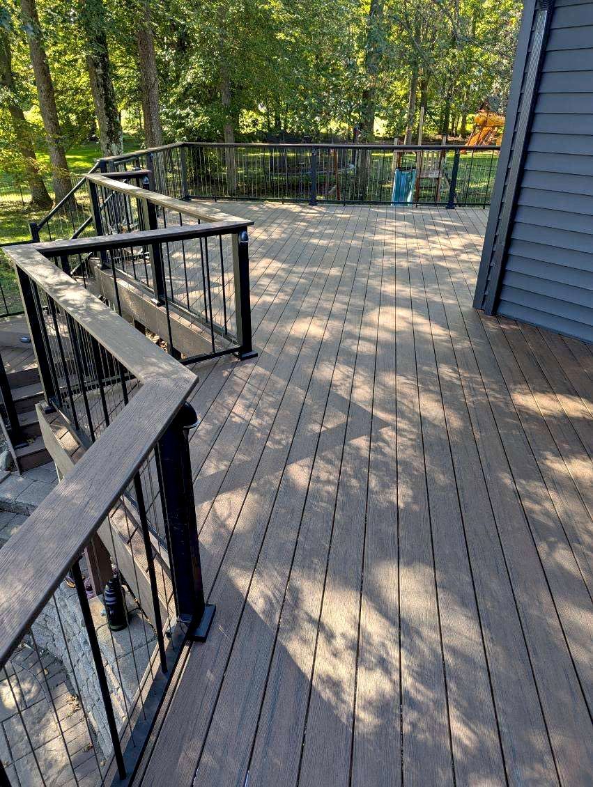 Wooden deck with black railing in a sunlit wooded area, with blue siding on the right