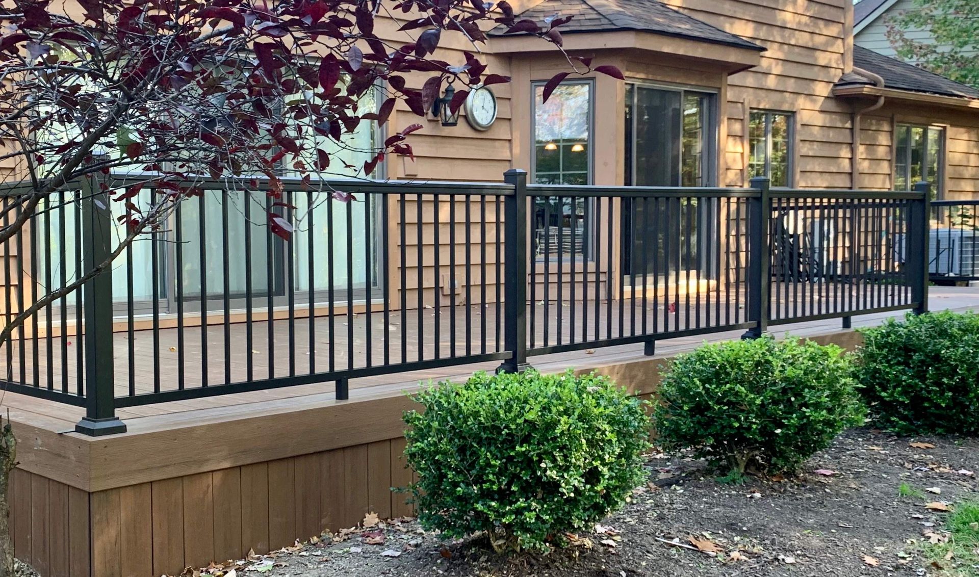 Black metal railing on a wooden deck beside a house, with shrubs and a tree in the foreground