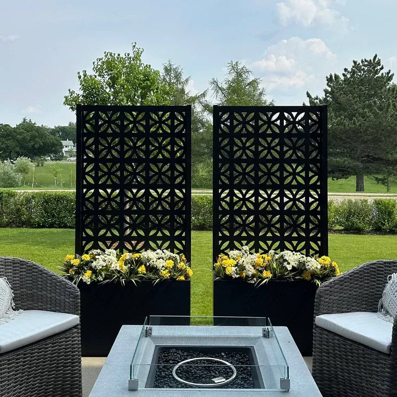 Patio lounge with two black lattice screens, flower boxes, and white woven chairs around a fire table.