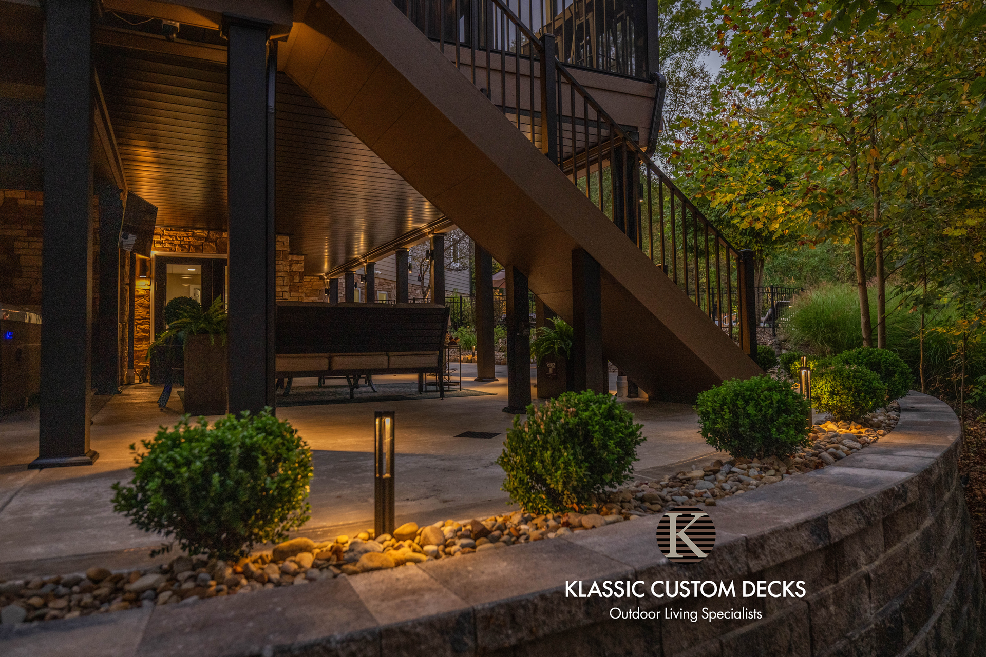 Outdoor patio with stone steps, staircase, shrubs, and warm evening lighting beside a house