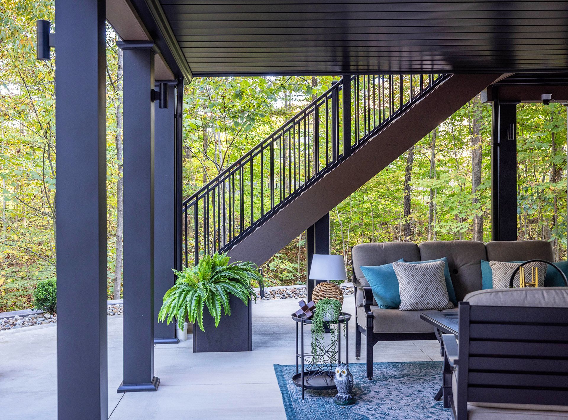 Covered patio with black staircase, gray sofa, potted plant, and wooded backyard view