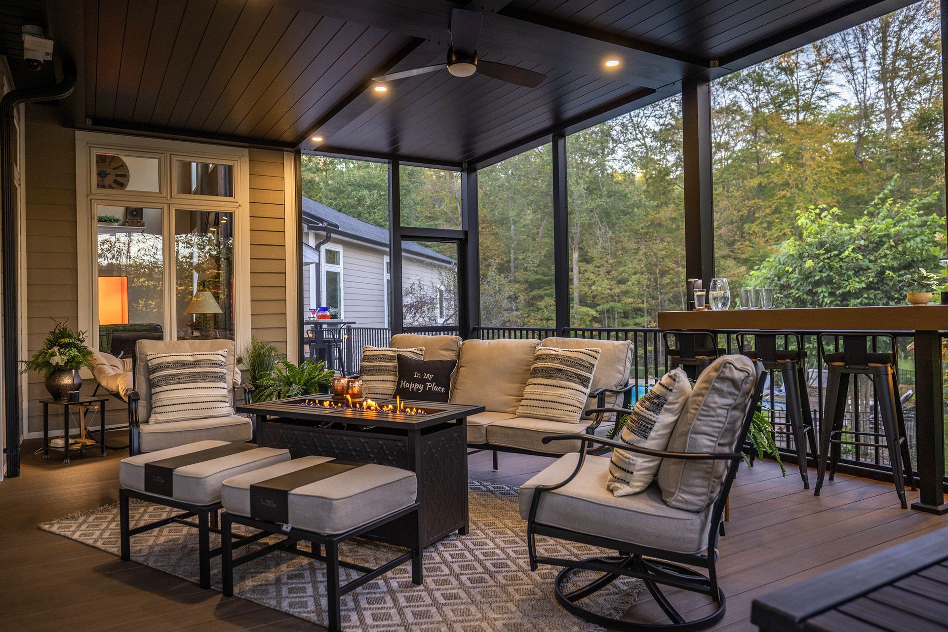 Covered patio with wicker seating, fire pit, and bar stools overlooking a wooded yard
