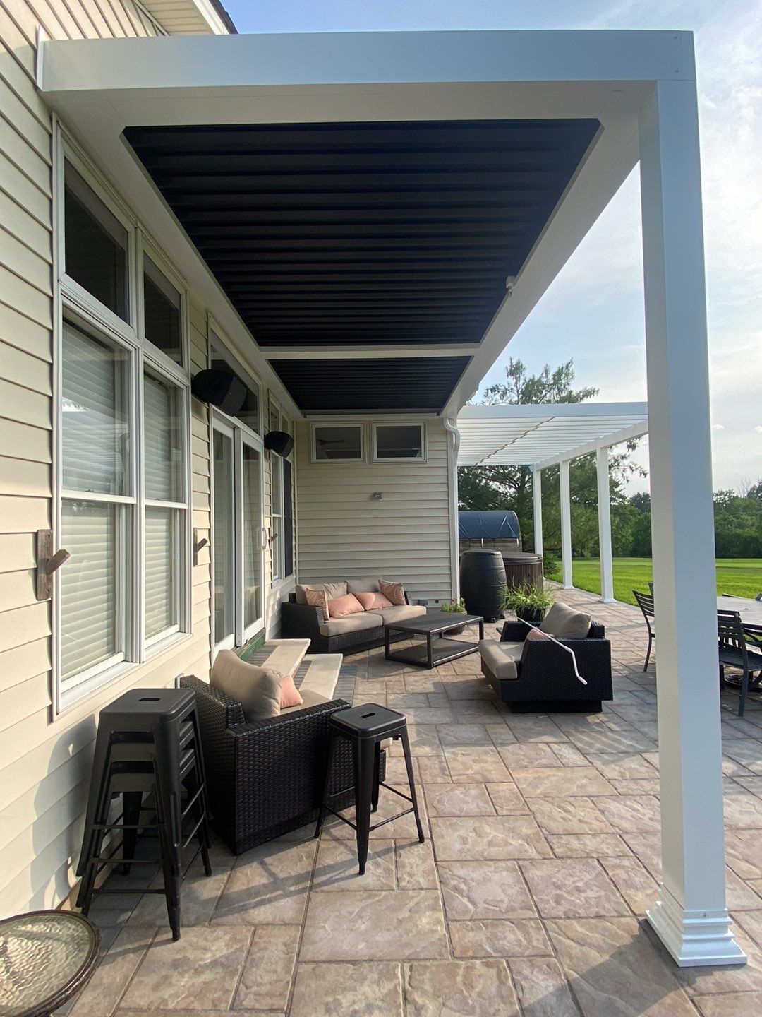 Covered patio with wicker seating, cushions, and stone pavers beside a house, overlooking a grassy yard