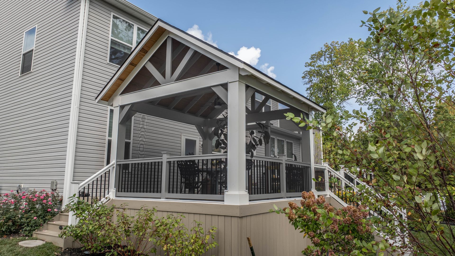 Covered porch with gray railings and wooden trusses on a light gray house, surrounded by shrubs and trees