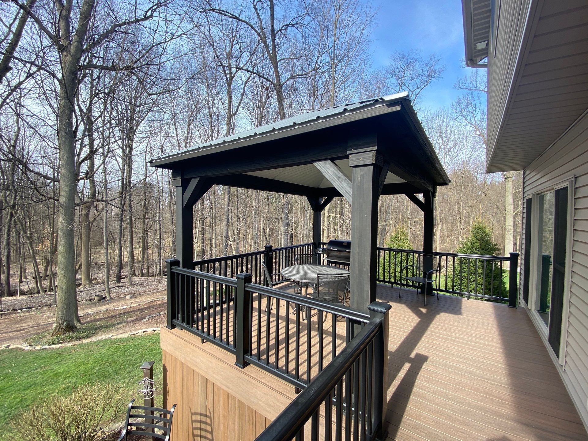 Covered wooden deck with black railing, gazebo, and seating beside a house in a wooded yard.