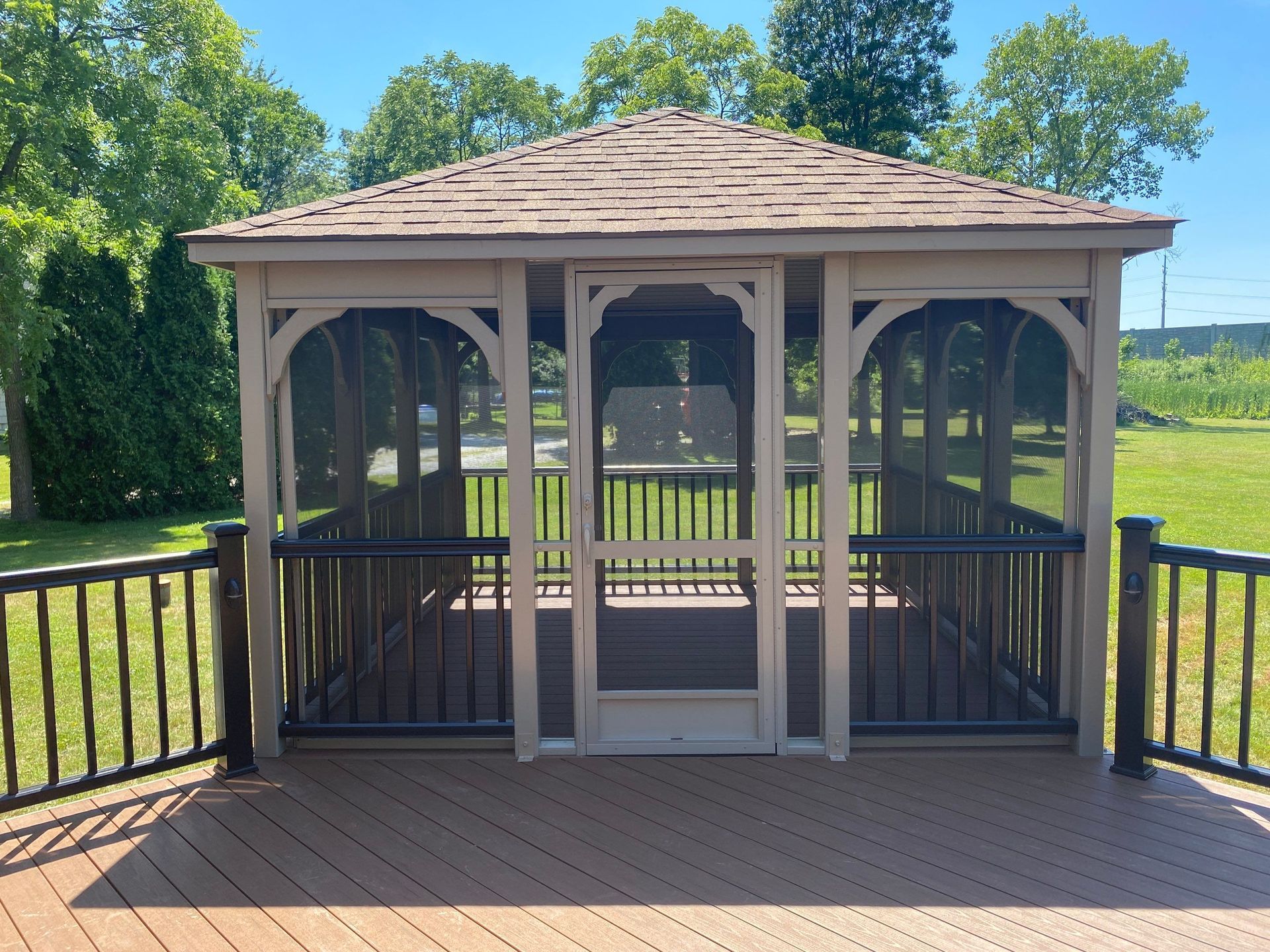 Wooden gazebo with screened sides on a deck in a sunny grassy yard.