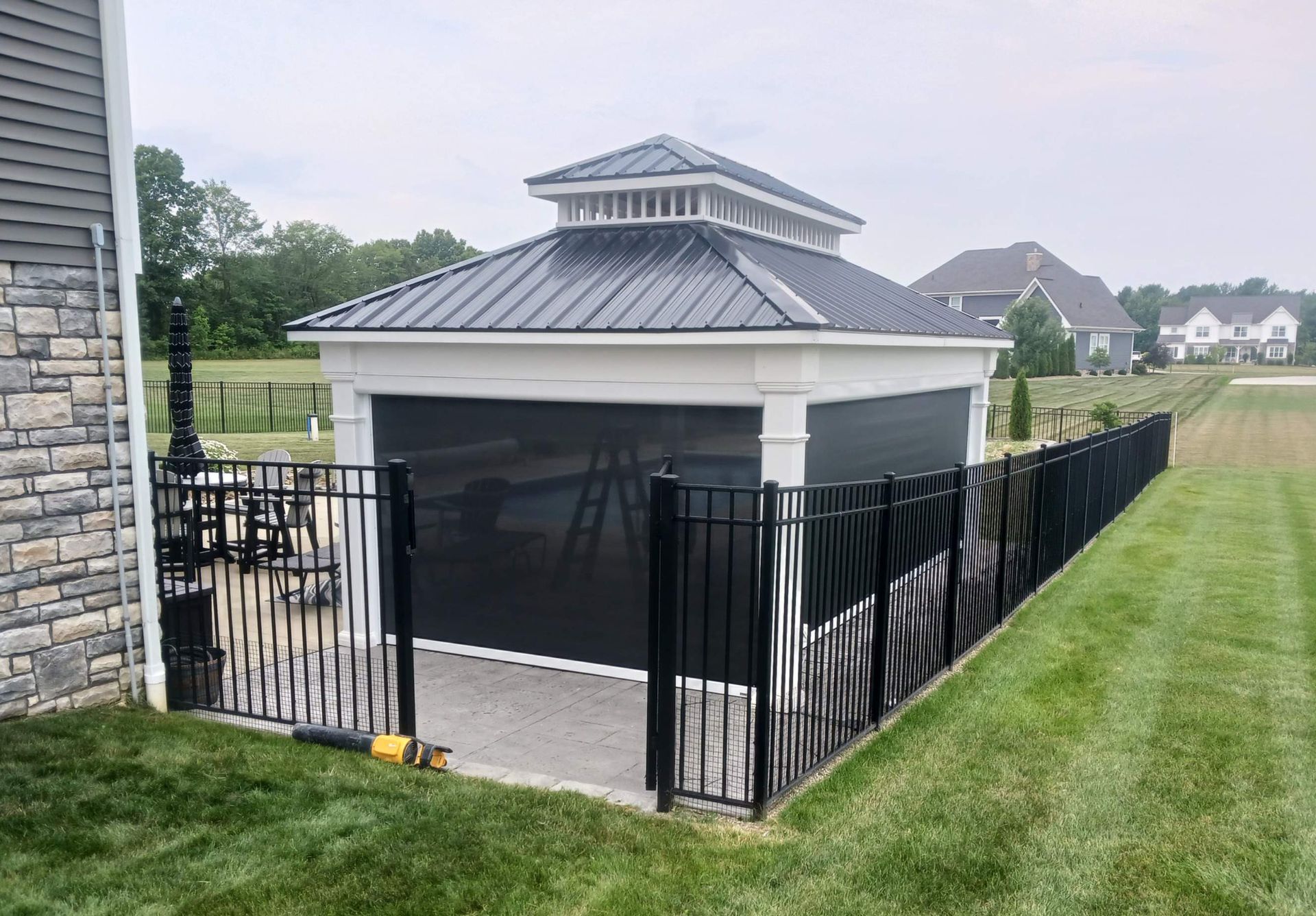 Black-fenced backyard pool enclosure beside a house, with a grassy lawn and neighboring homes nearby