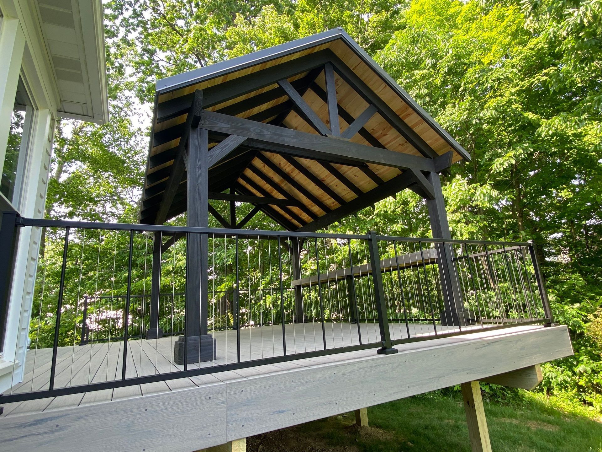 Covered wooden pavilion with black railings on a raised deck in a leafy backyard