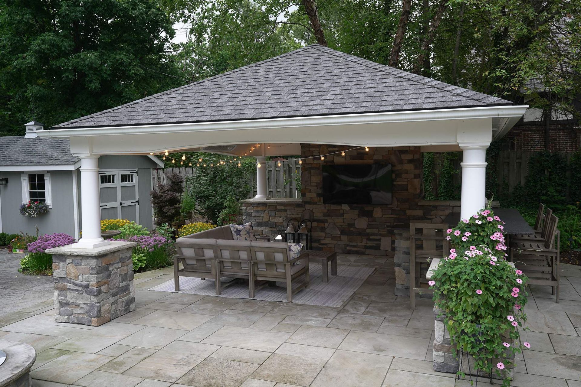 Stone patio gazebo with white columns, wicker chairs, and hanging flowers in a garden setting