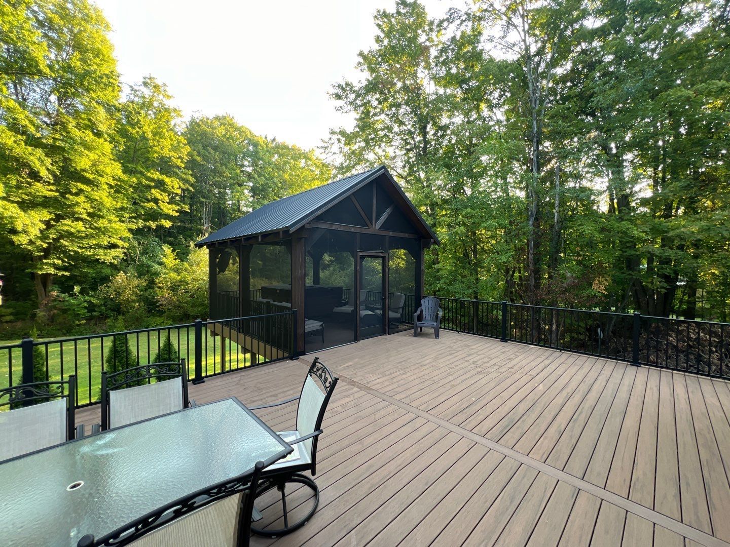 Outdoor deck with glass table and chairs beside a small black cabin in a wooded area