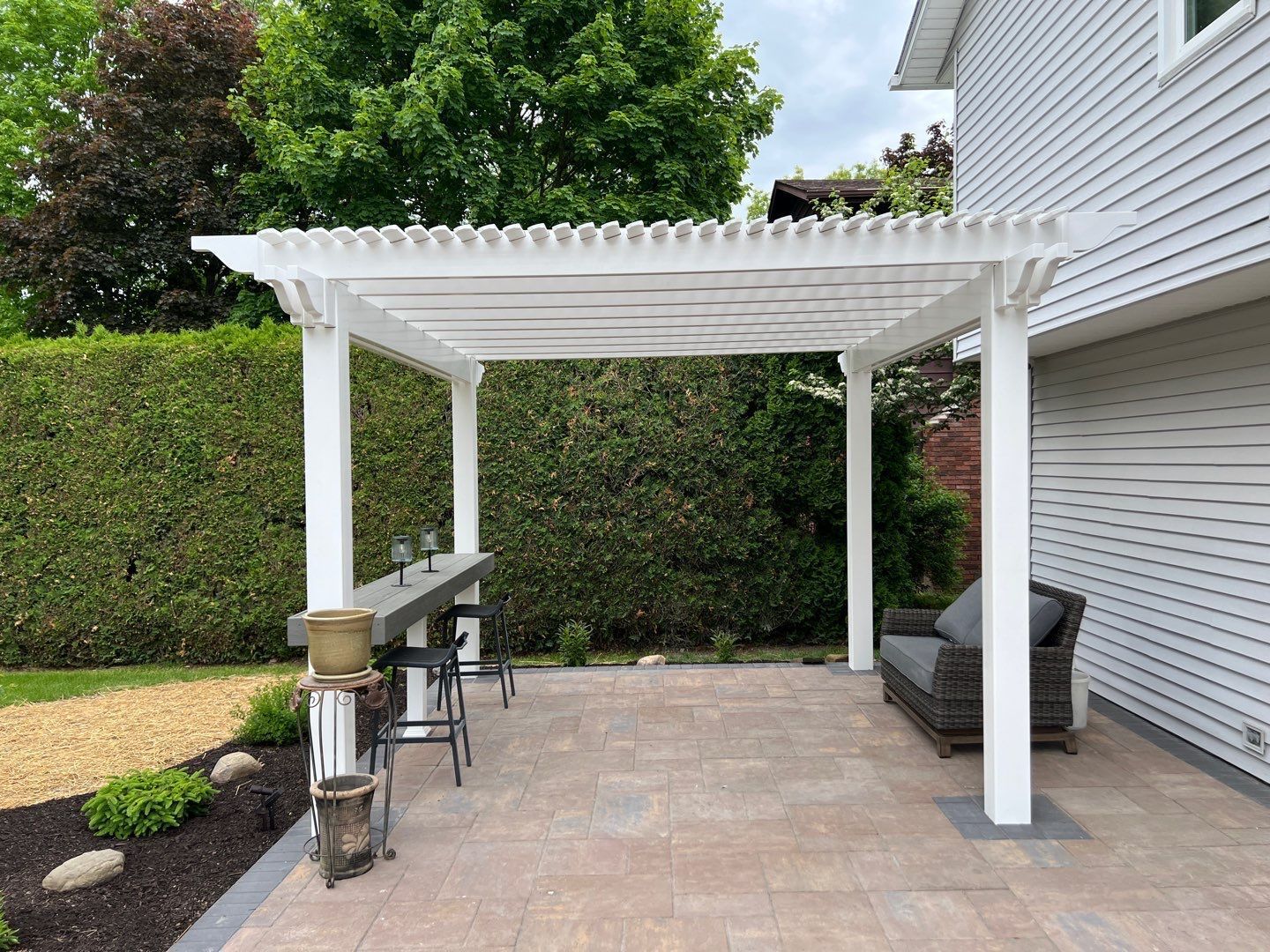 White pergola over a paved patio beside a house, with a grill and outdoor seating by a hedge.