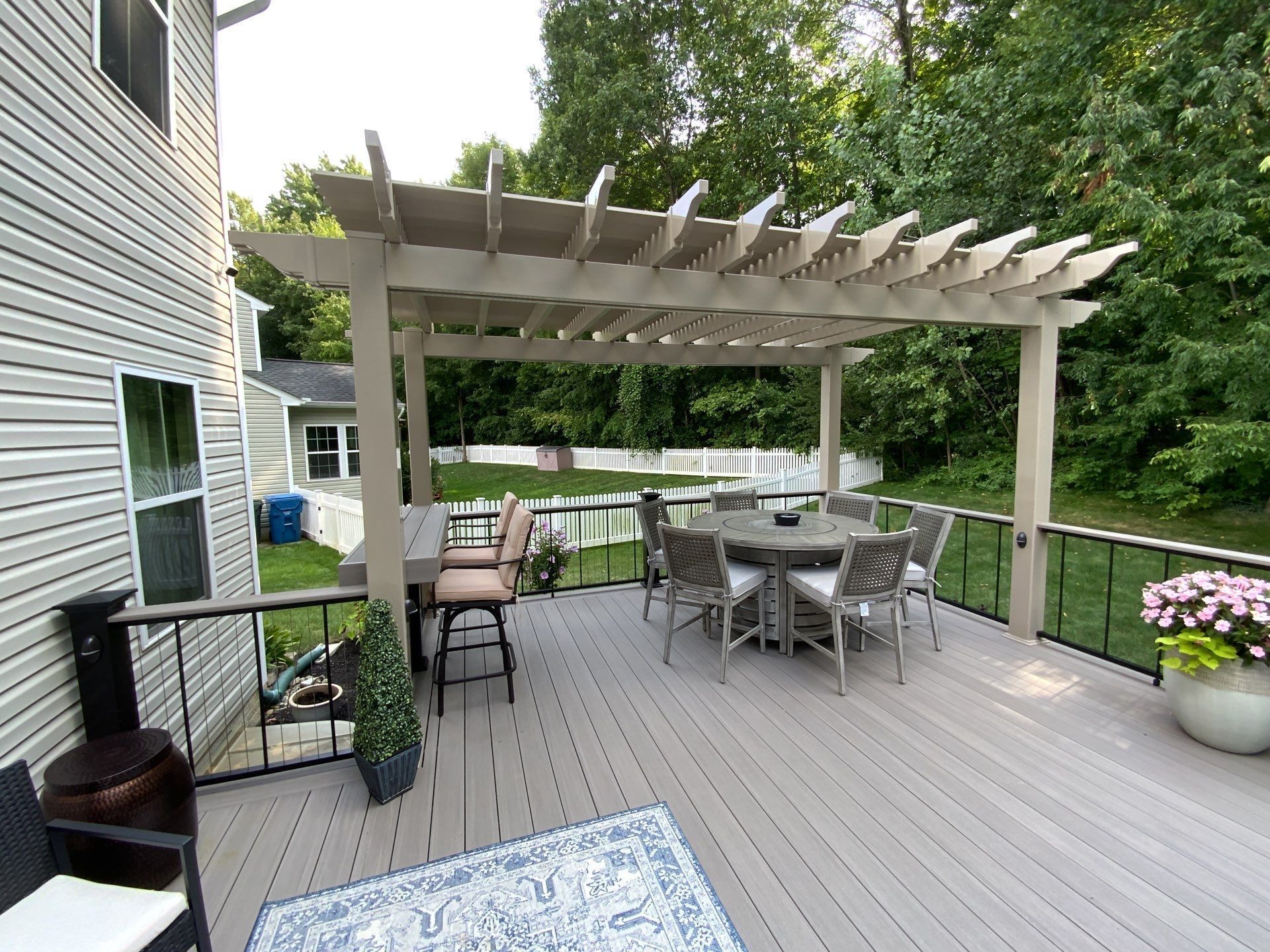 Backyard deck with pergola, outdoor dining table, chairs, grill, and potted flowers by a house