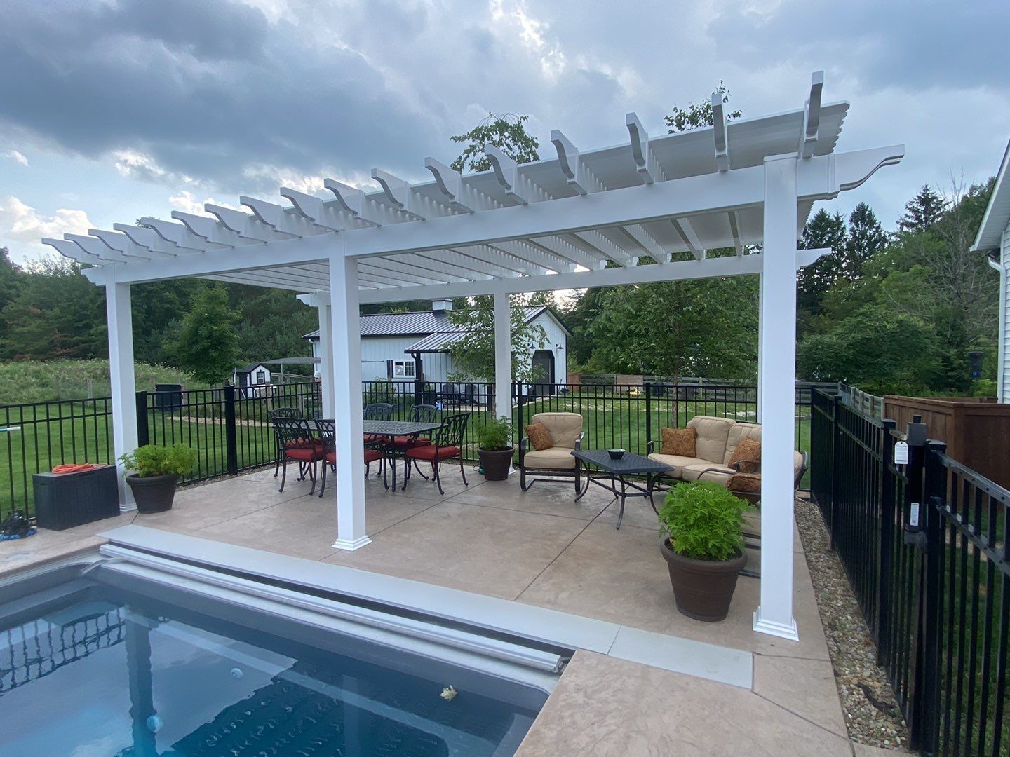 White pergola beside a pool with patio seating, potted plants, and a black fence in a backyard garden
