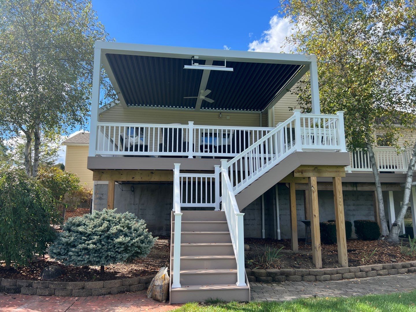 Elevated white deck with stairs and railing on a house, framed by trees and blue sky