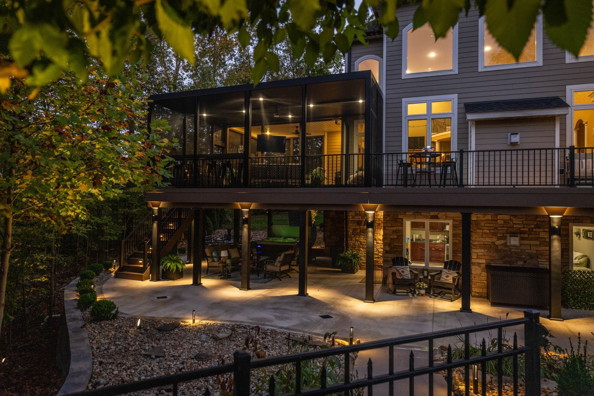 Modern house with lit patio and glass-enclosed deck at dusk, surrounded by trees and landscaped garden
