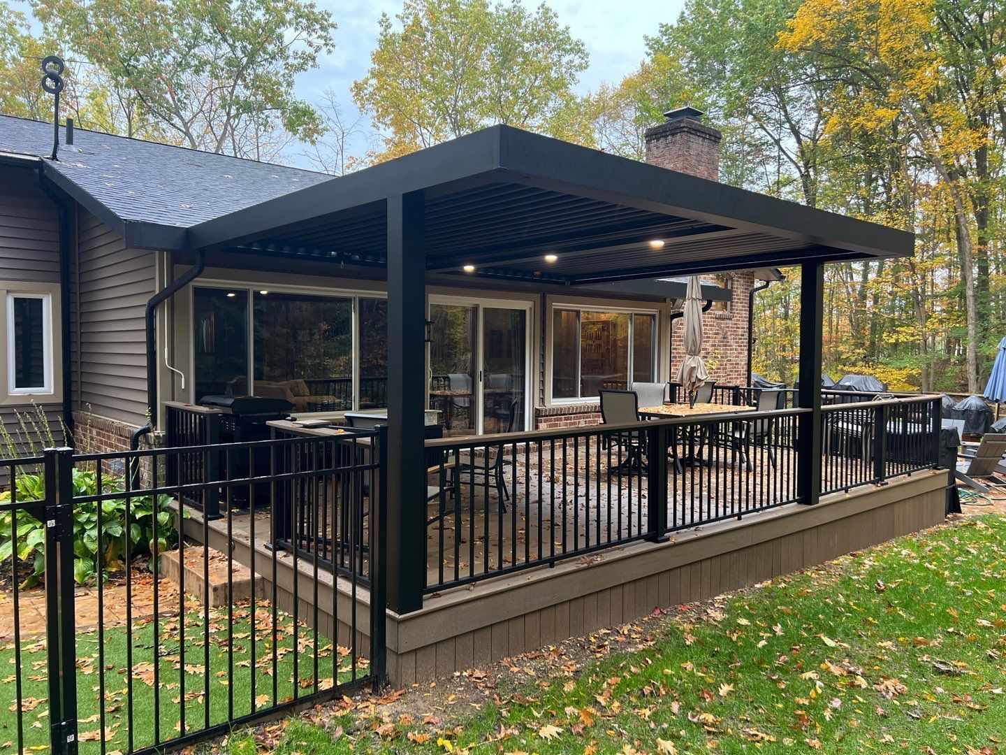 Covered backyard deck with black railings and outdoor seating beside a house, surrounded by autumn trees