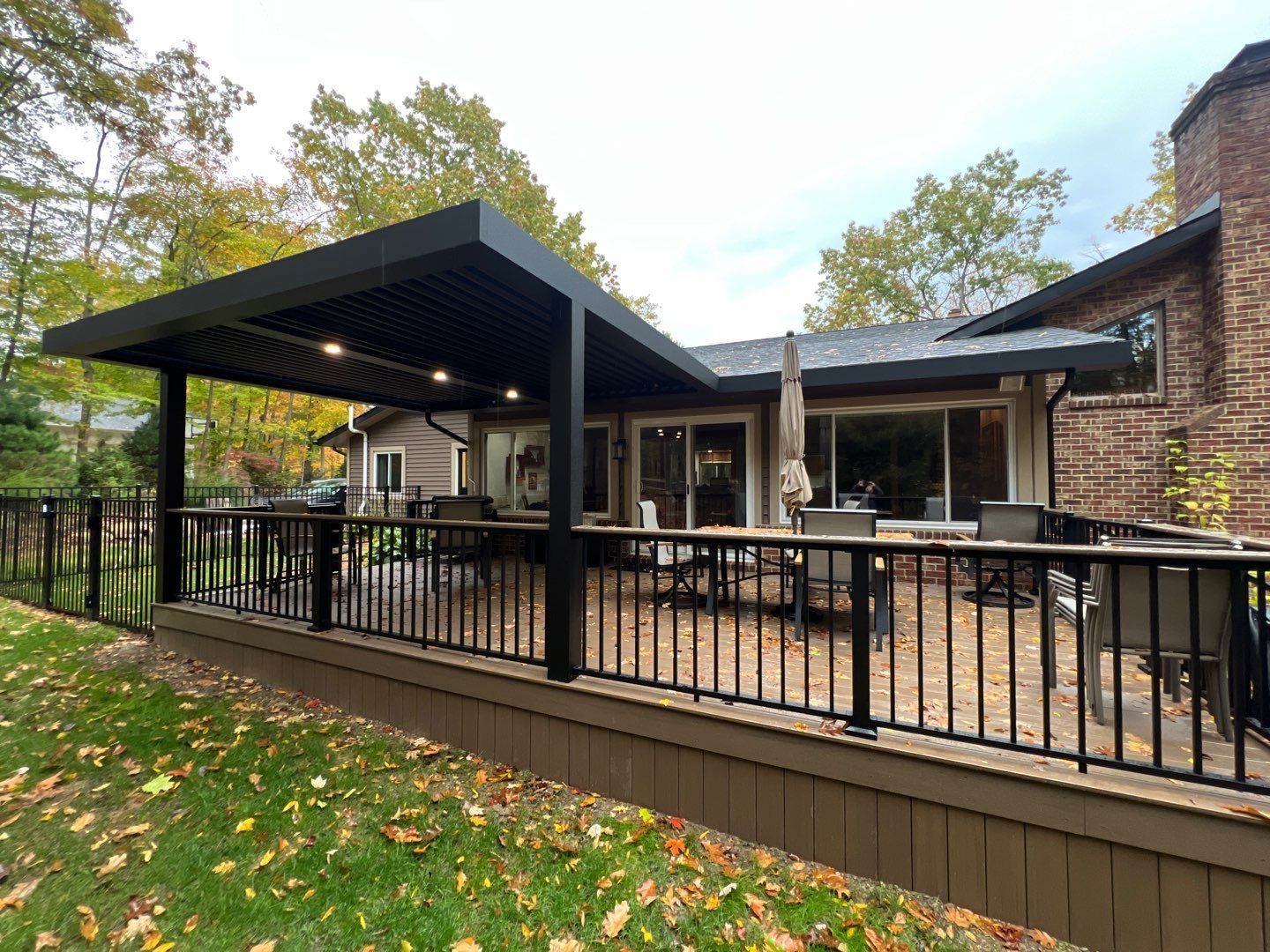 Backyard deck with black pergola, railing, and outdoor seating beside a brick house in autumn