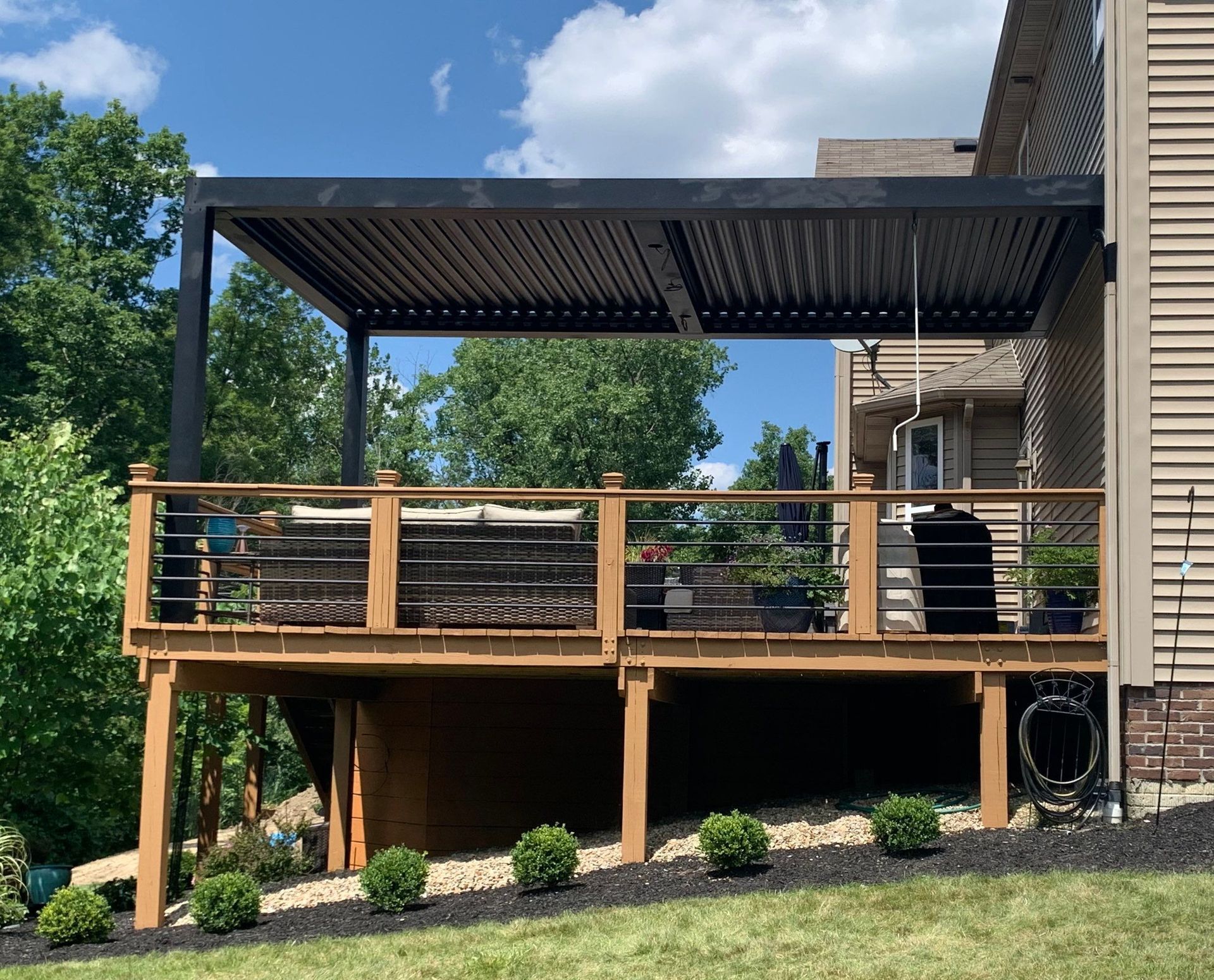 Wooden backyard deck with black pergola and railing beside a house, under a blue sky