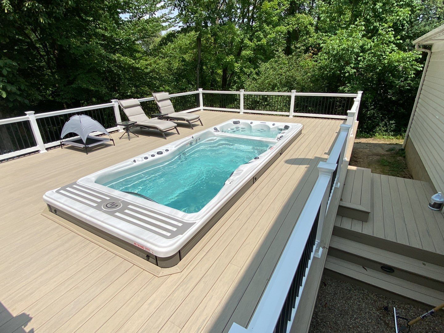 Raised deck with a rectangular above-ground pool, lounge chairs, and white railing surrounded by trees.