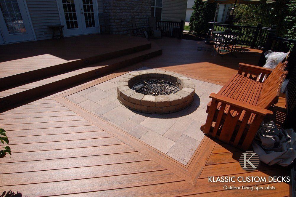 Wooden deck with a circular stone fire pit and benches in a sunny backyard patio