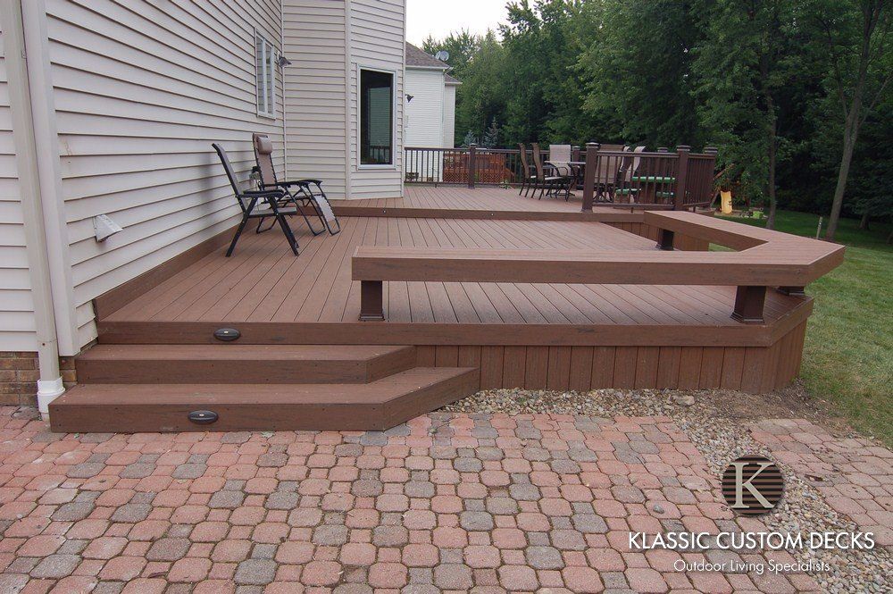 Raised wooden deck with steps and bench seating beside a house, bordered by a brick patio.