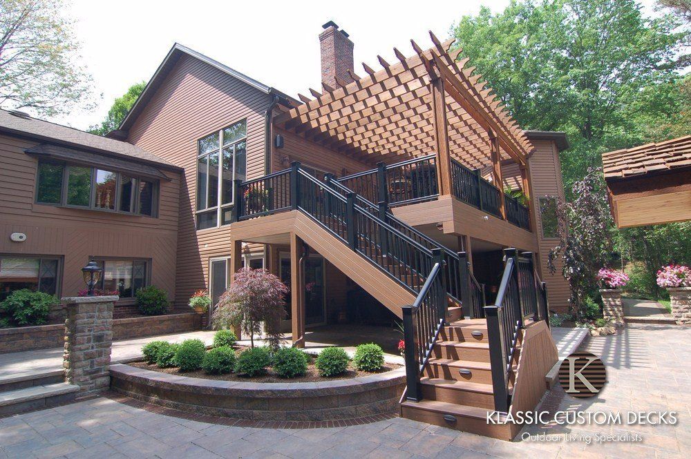 Brick home with a large wooden deck, stairs, and pergola in a landscaped courtyard.