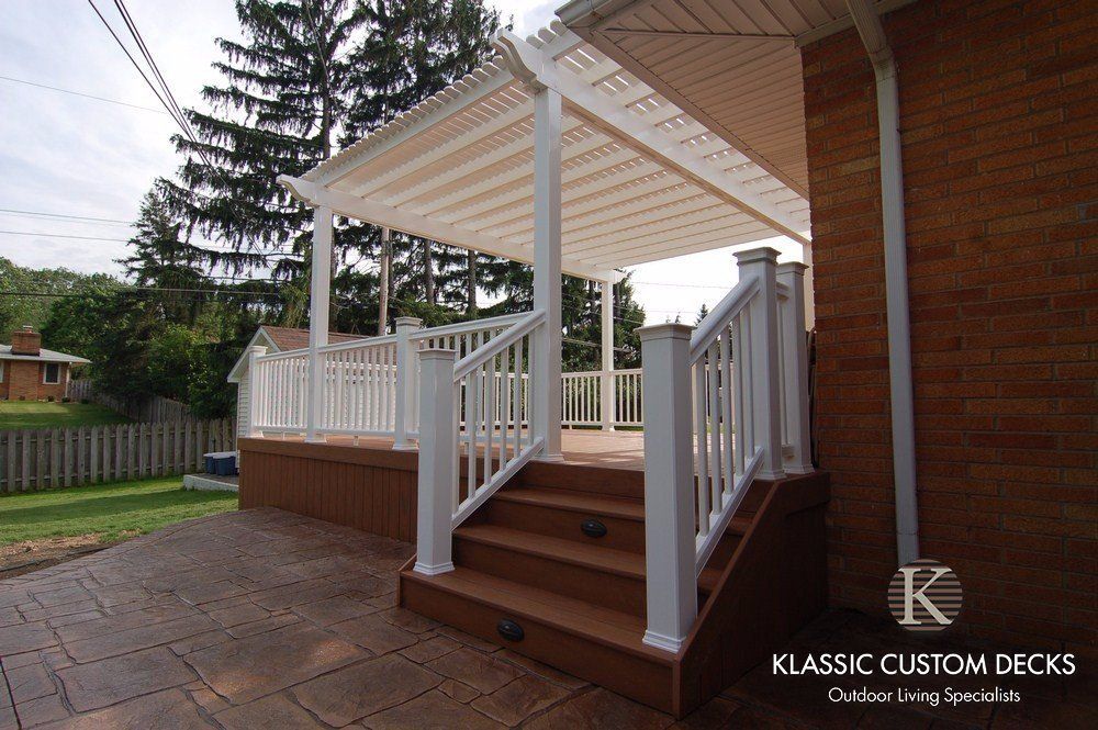 White pergola-covered wooden deck with stairs and railings beside a brick house, in a backyard garden