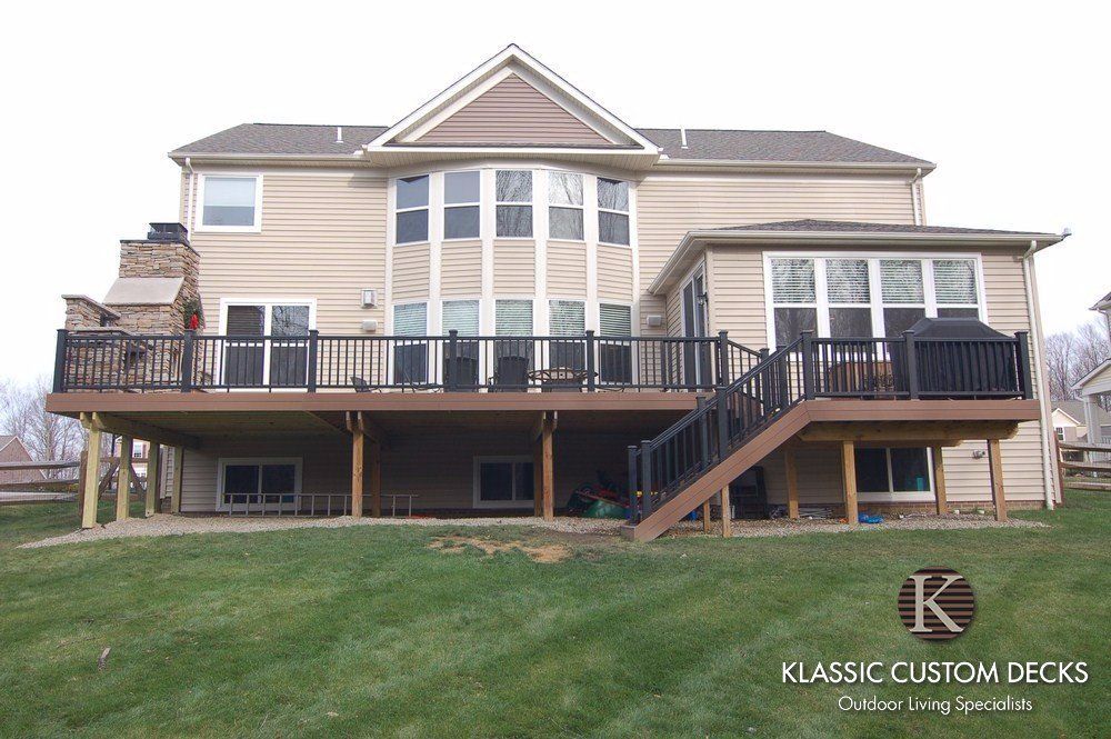 Large beige house with a raised wooden deck, black railings, and stairs in a grassy yard.
