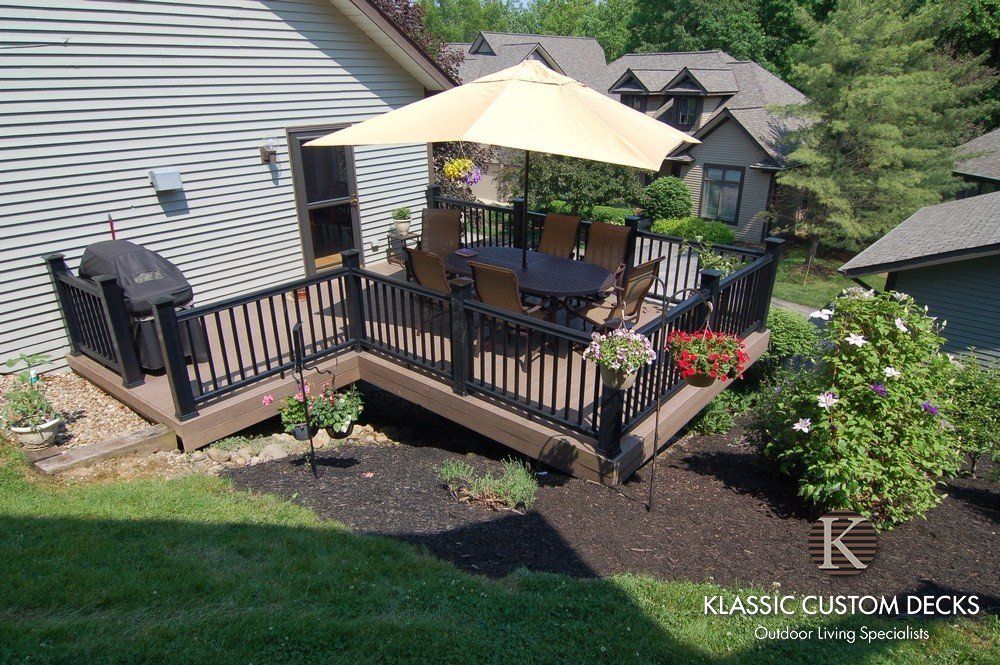 Raised wooden deck with black railings, beige umbrella, grill, and potted flowers beside a house