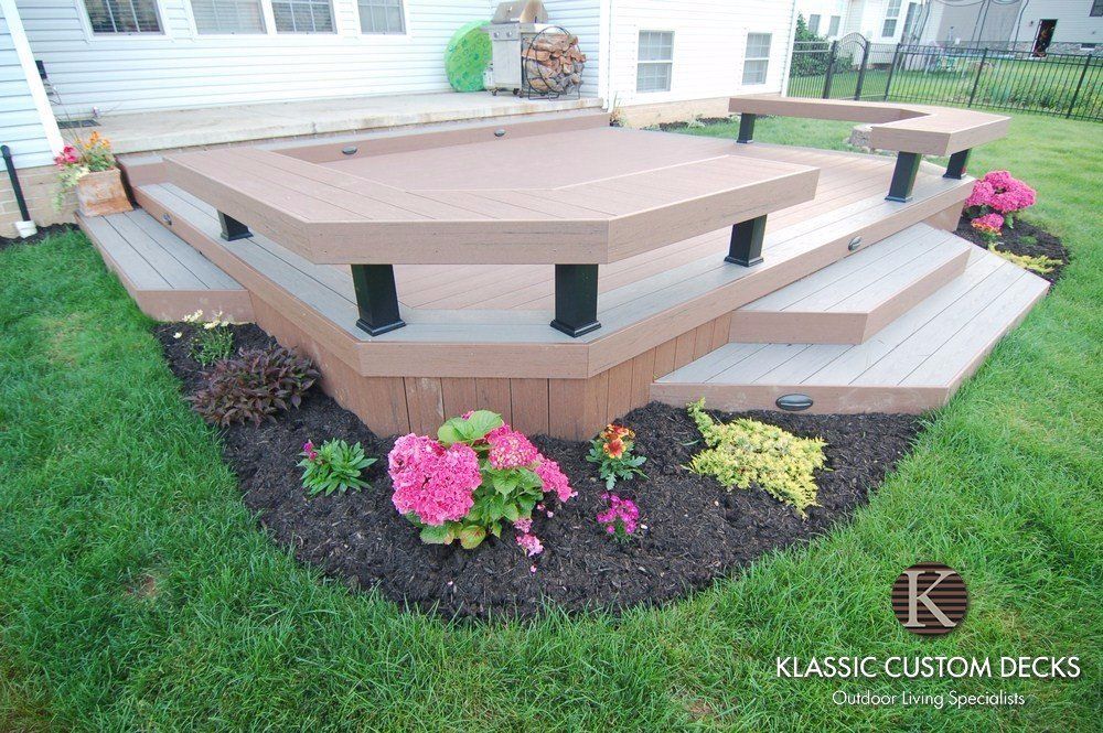 Raised octagonal wooden deck with beige railings and steps, surrounded by grass and pink flowers by a house