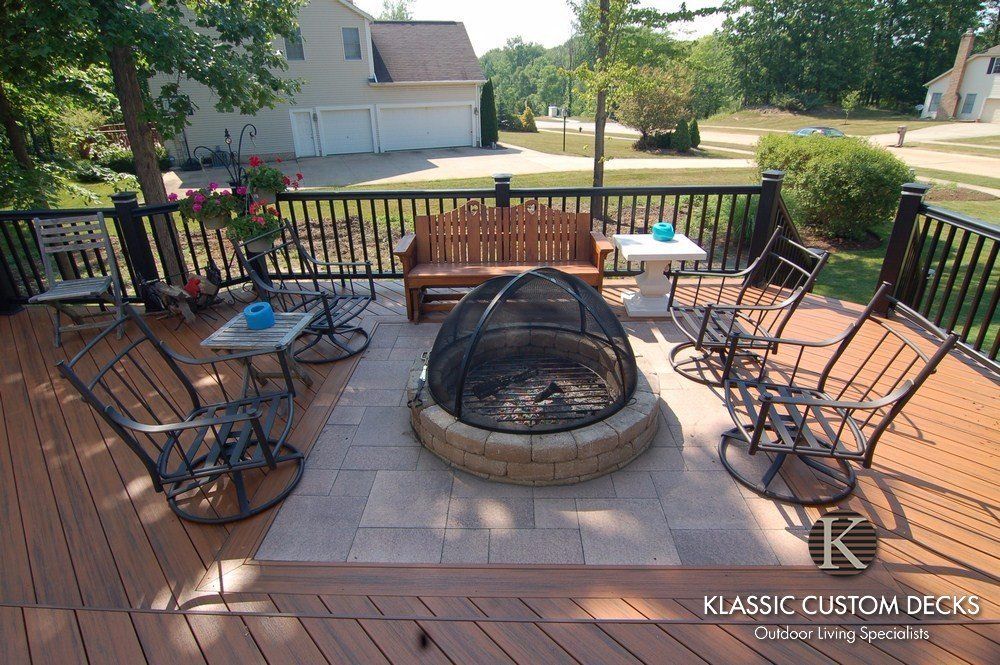 Backyard deck with cushioned chairs around a circular fire pit and a glass-top table, overlooking a lawn