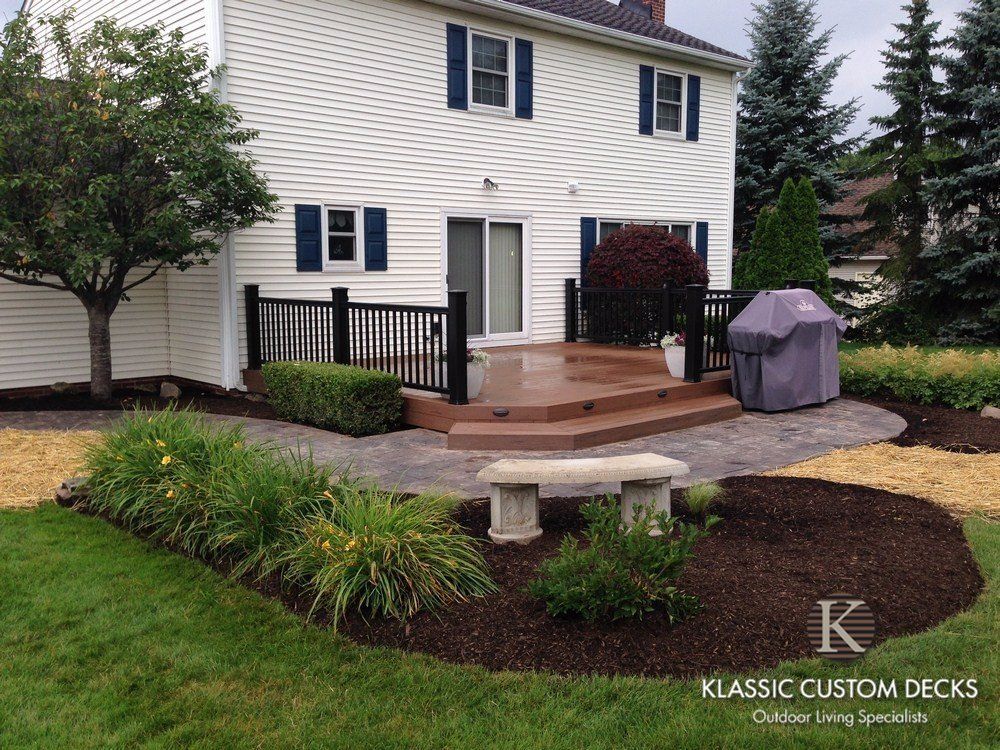 Landscaped backyard patio with steps, black railing, and a covered grill beside a white house