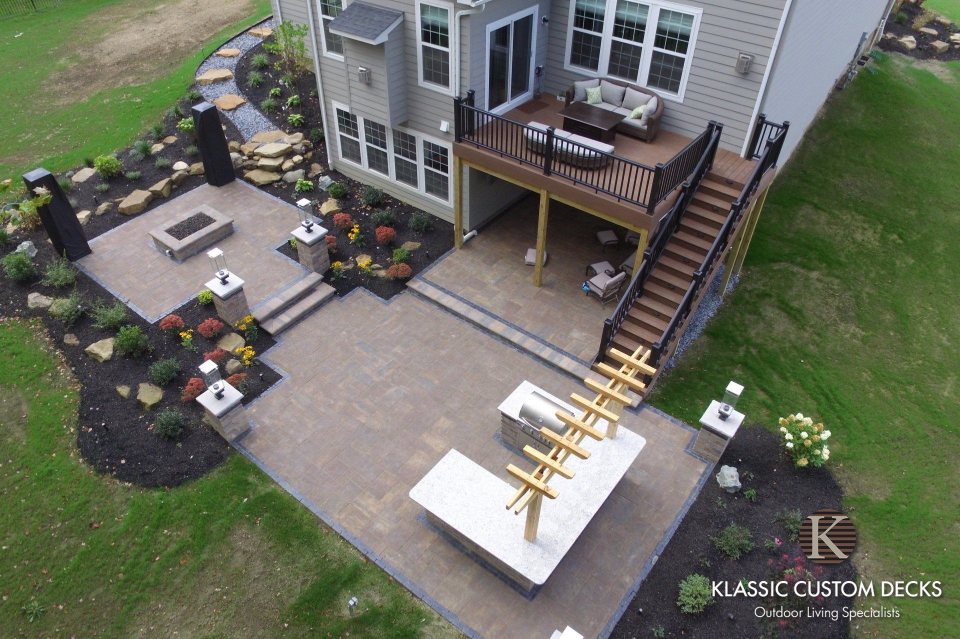 Aerial view of a house patio with paver walkway, stone fire pit area, deck, and stairs over a landscaped yard