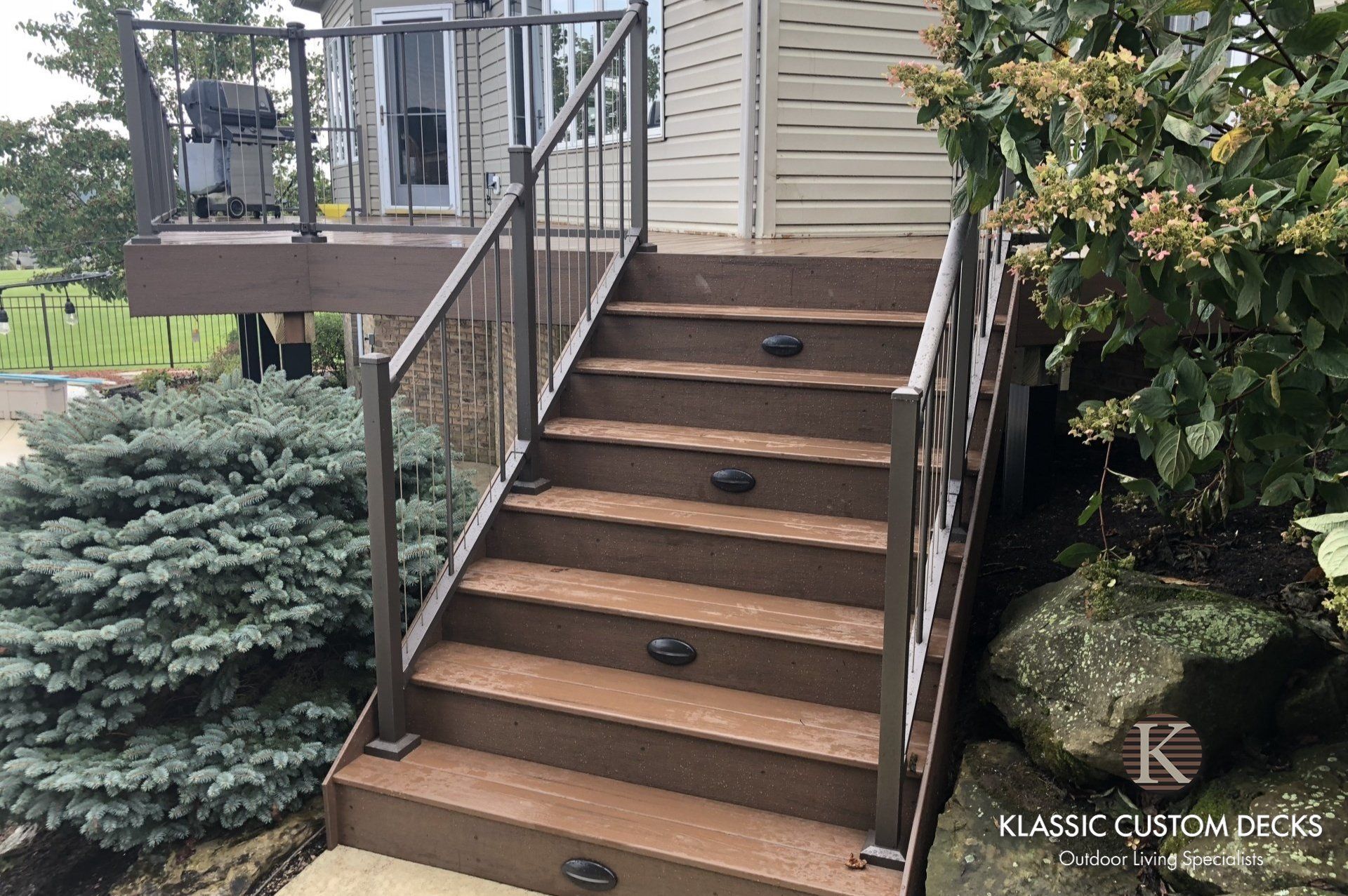 Front stairs with brown steps and metal railings leading to a house entry, bordered by shrubs and flowers
