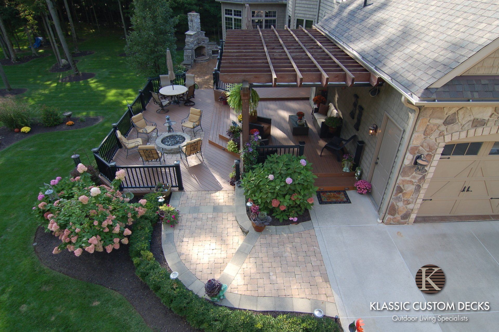 Backyard patio with pergola, brick walkway, fountain, benches, and flowering shrubs beside a house