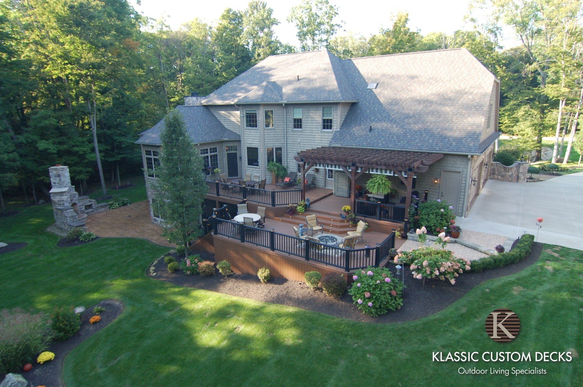 Aerial view of a large stone house with a wooden deck, patio seating, and landscaped backyard