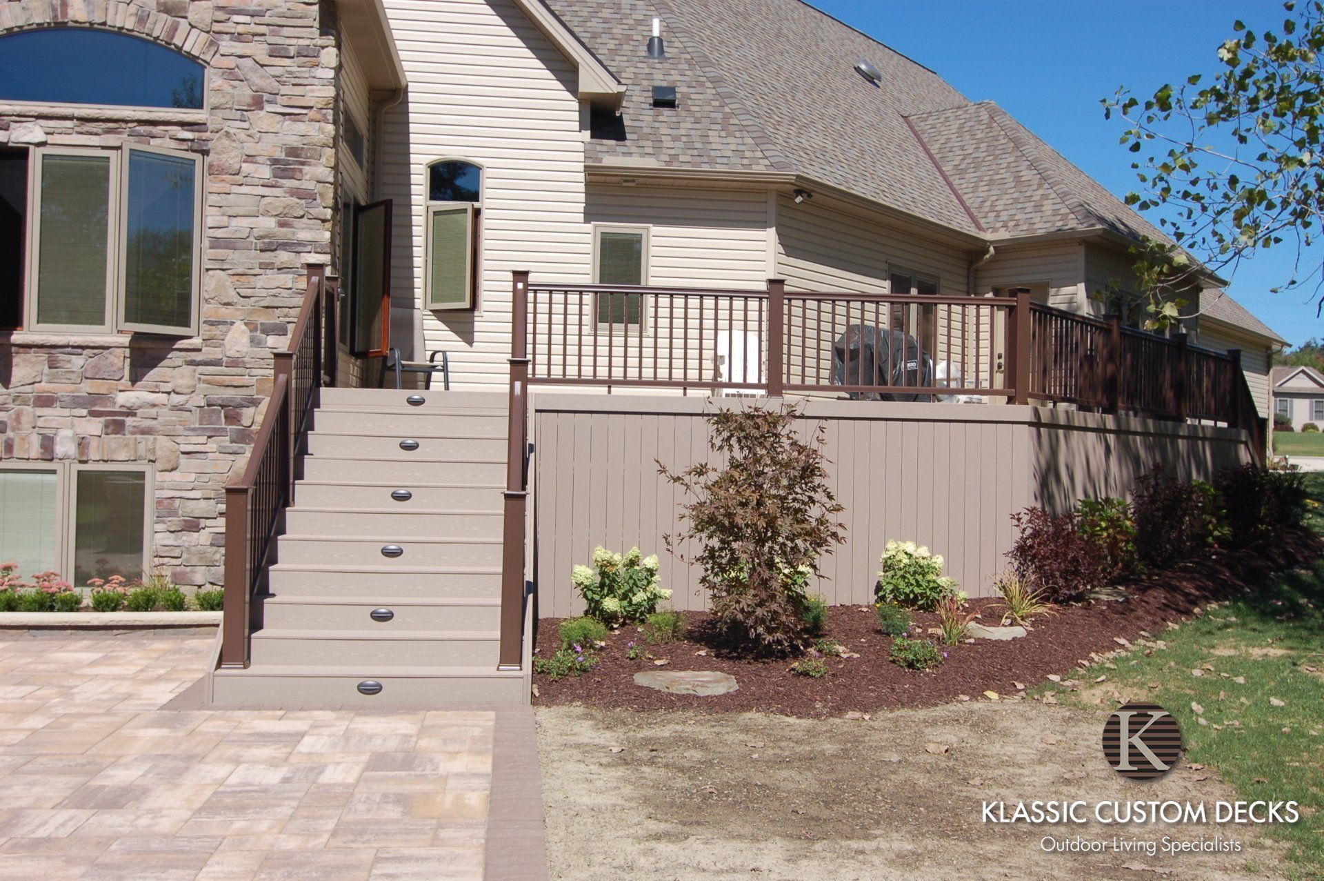 Stone house exterior with front staircase, tan deck railing, and landscaped yard under a clear blue sky