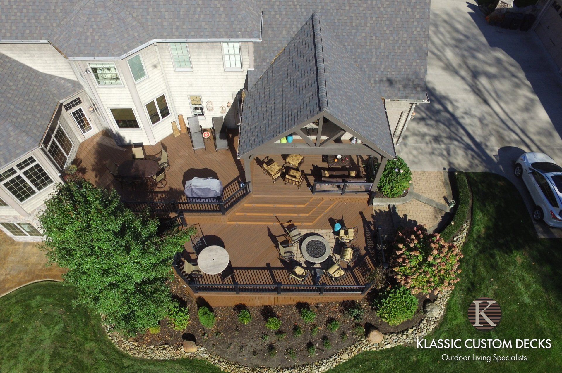 Aerial view of a backyard deck with tables, chairs, lounge seating, and a covered pergola beside a house.