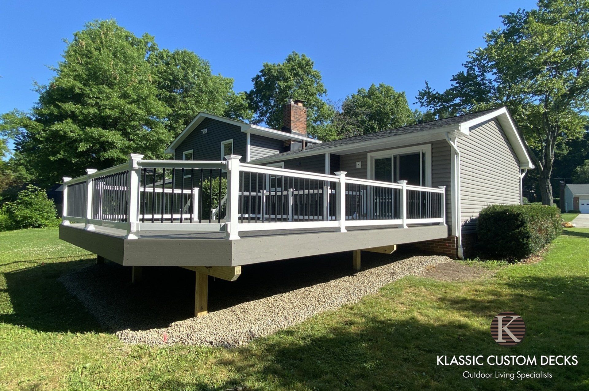 White deck with railings attached to a house in a sunny yard, with trees and grass around it