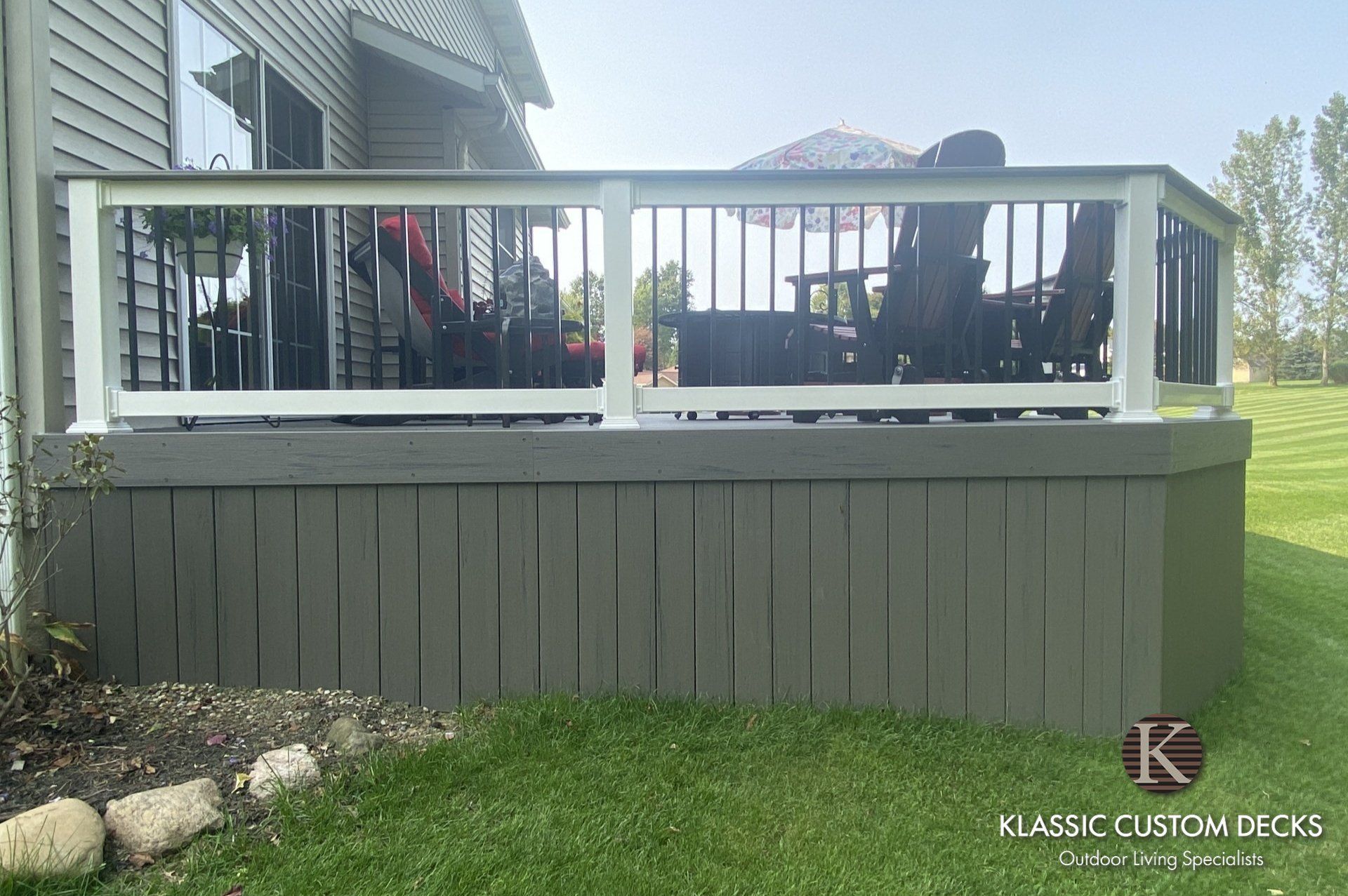 Raised deck with white railing and gray skirting beside a house, with patio chairs and grass below.