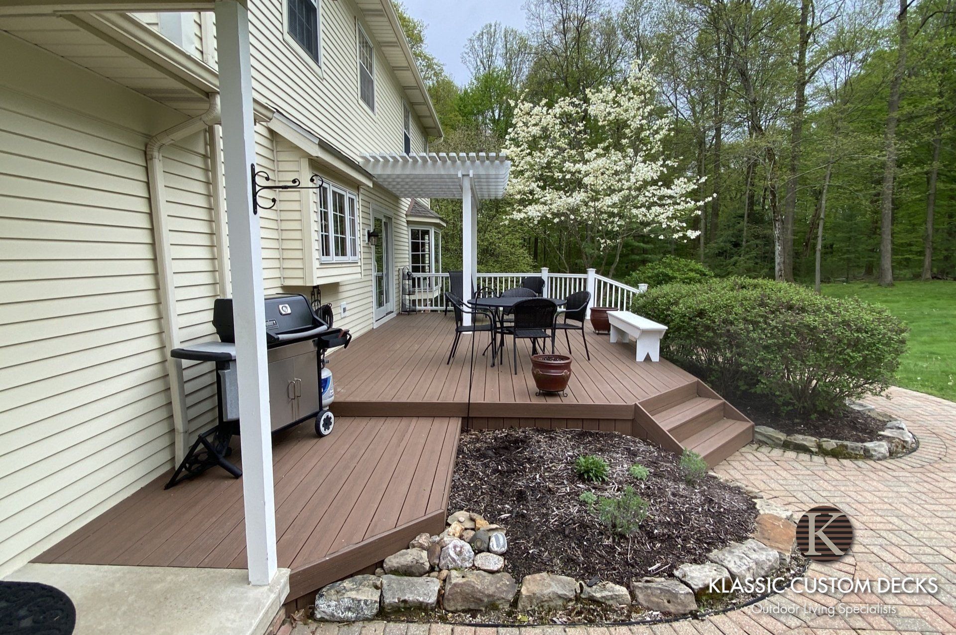 Backyard deck with grill, patio furniture, and steps beside a beige house and spring trees