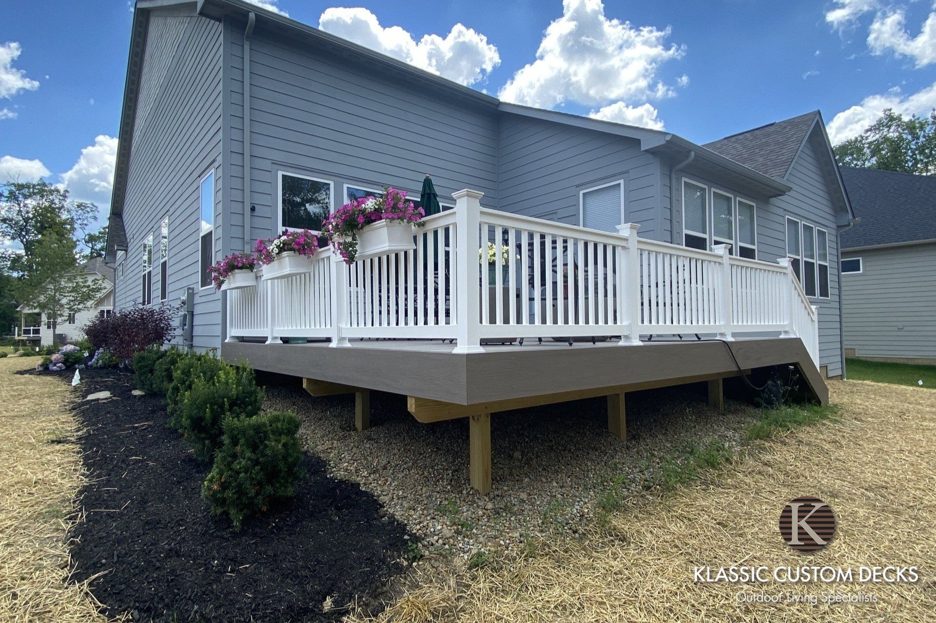 Back deck with white railing on a gray house, set above a sloped yard under a blue sky.