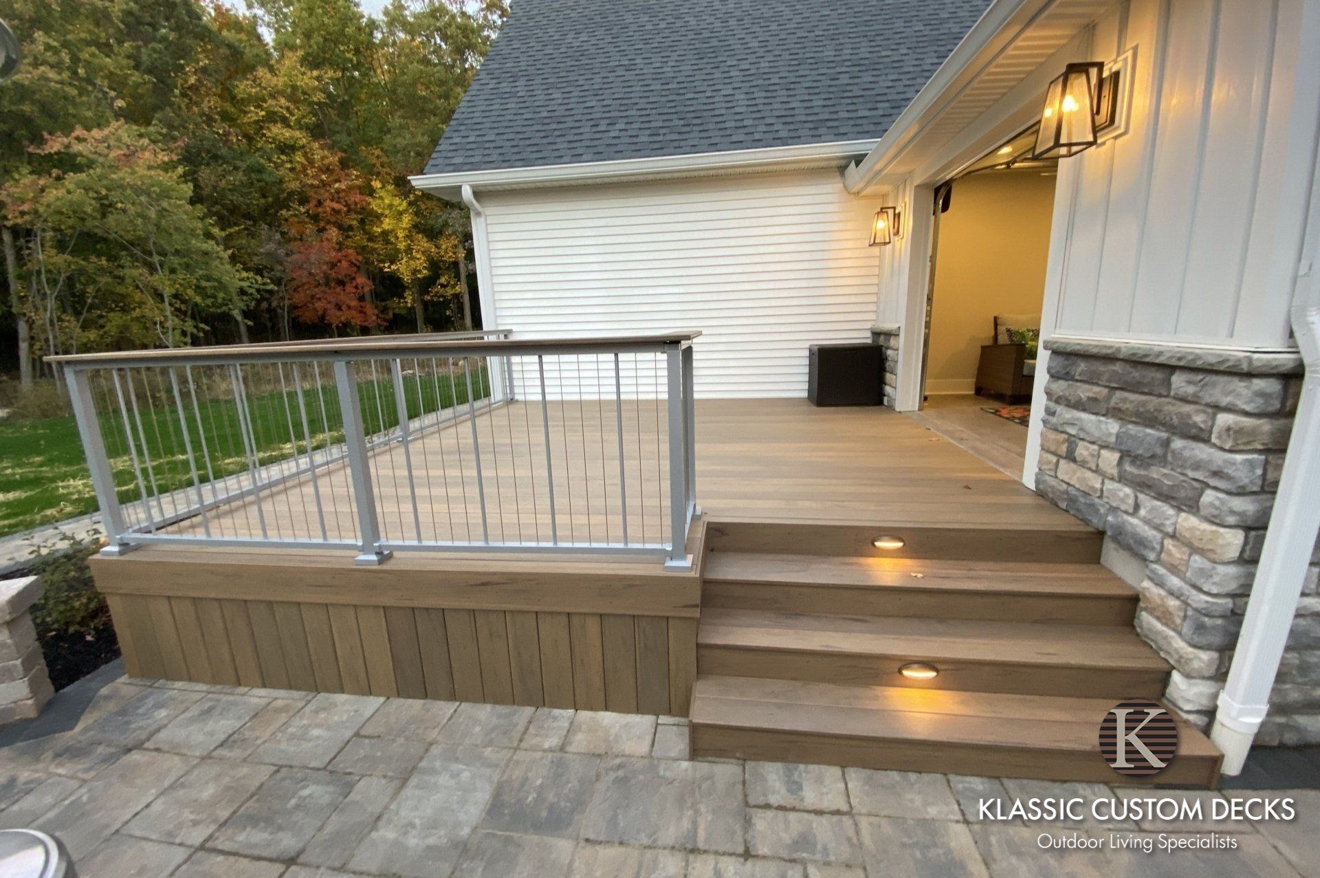 Backyard deck with steps, railing, and stone patio beside a white house at dusk